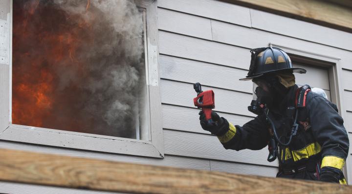 Firefighter holding thermal imaging camera up to a window with smoke coming out