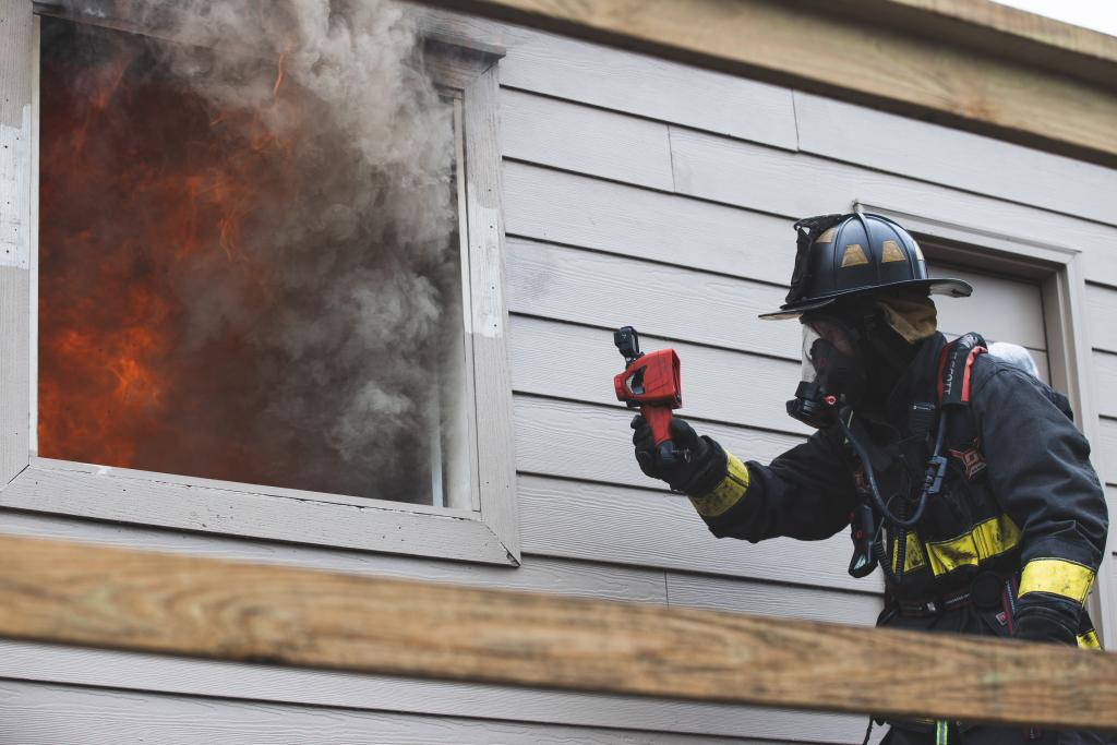 Firefighter holding thermal imaging camera up to a window with smoke coming out