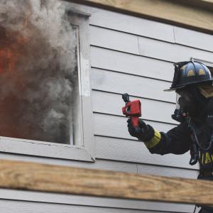 Firefighter holding thermal imaging camera up to a window with smoke coming out
