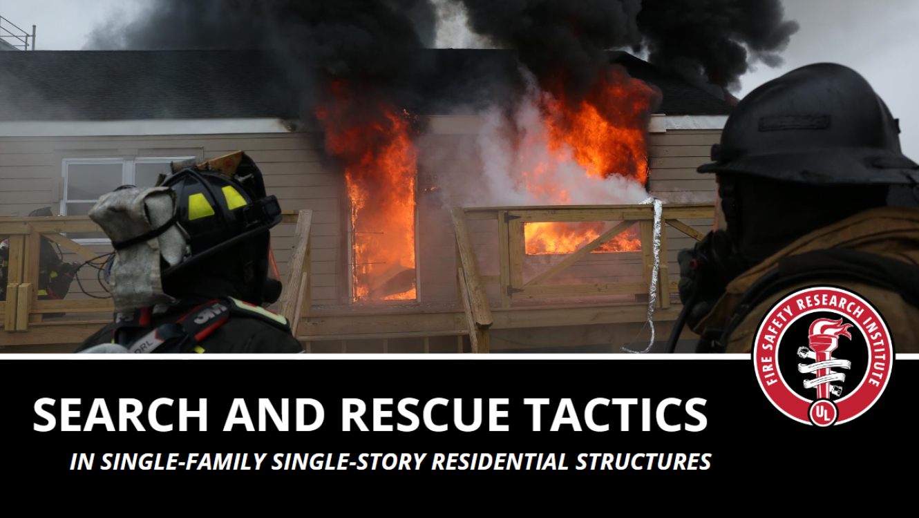 Two firefighters in black firefighter gear standing in front of a house with fire coming out of the left window